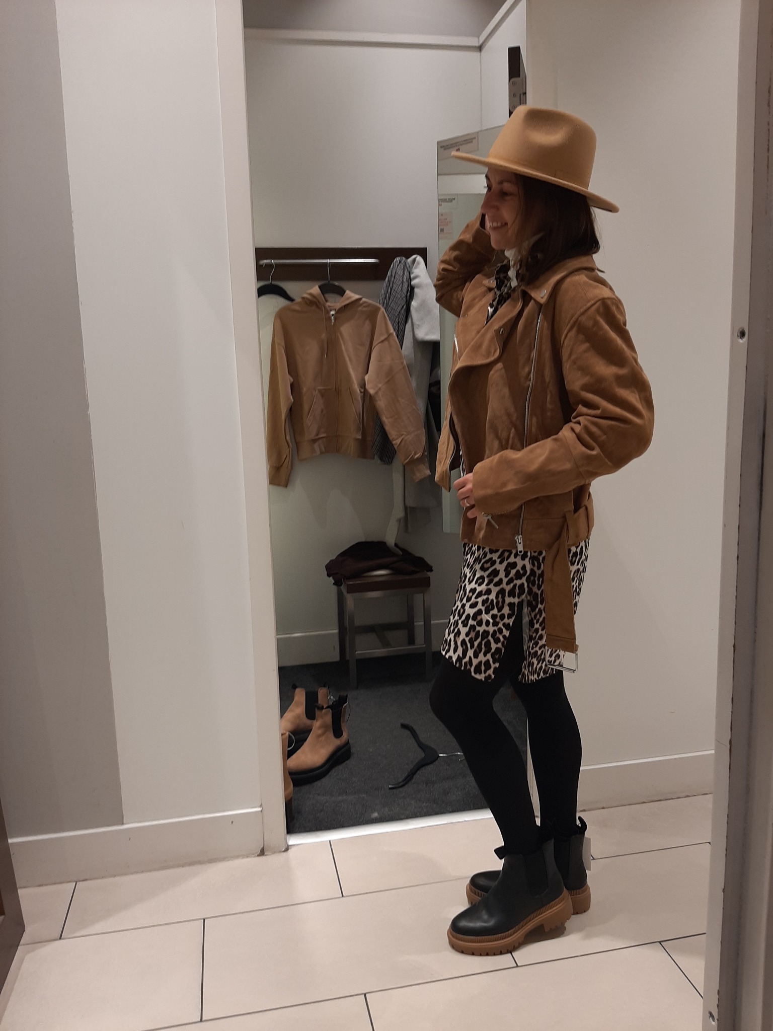 Kobieta uśmiecha się, przymierzając brązowy kapelusz i zamszową kurtkę w przymierzalni sklepu odzieżowego, widoczne czarne rajstopy i skórzane botki na grubej podeszwie.