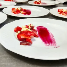 Przygotowane talerze z deserem: truskawki, biała czekolada i pur&eacute;e z owoc&oacute;w leśnych na białych talerzach z wytłaczanym wzorem, ustawione na metalowym stole.