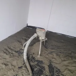 Aplikacja mokrej, szarej masy na czarnej folii podkładowej przy użyciu maszyny z wężem w rogu pomieszczenia, ściany w tle białe.