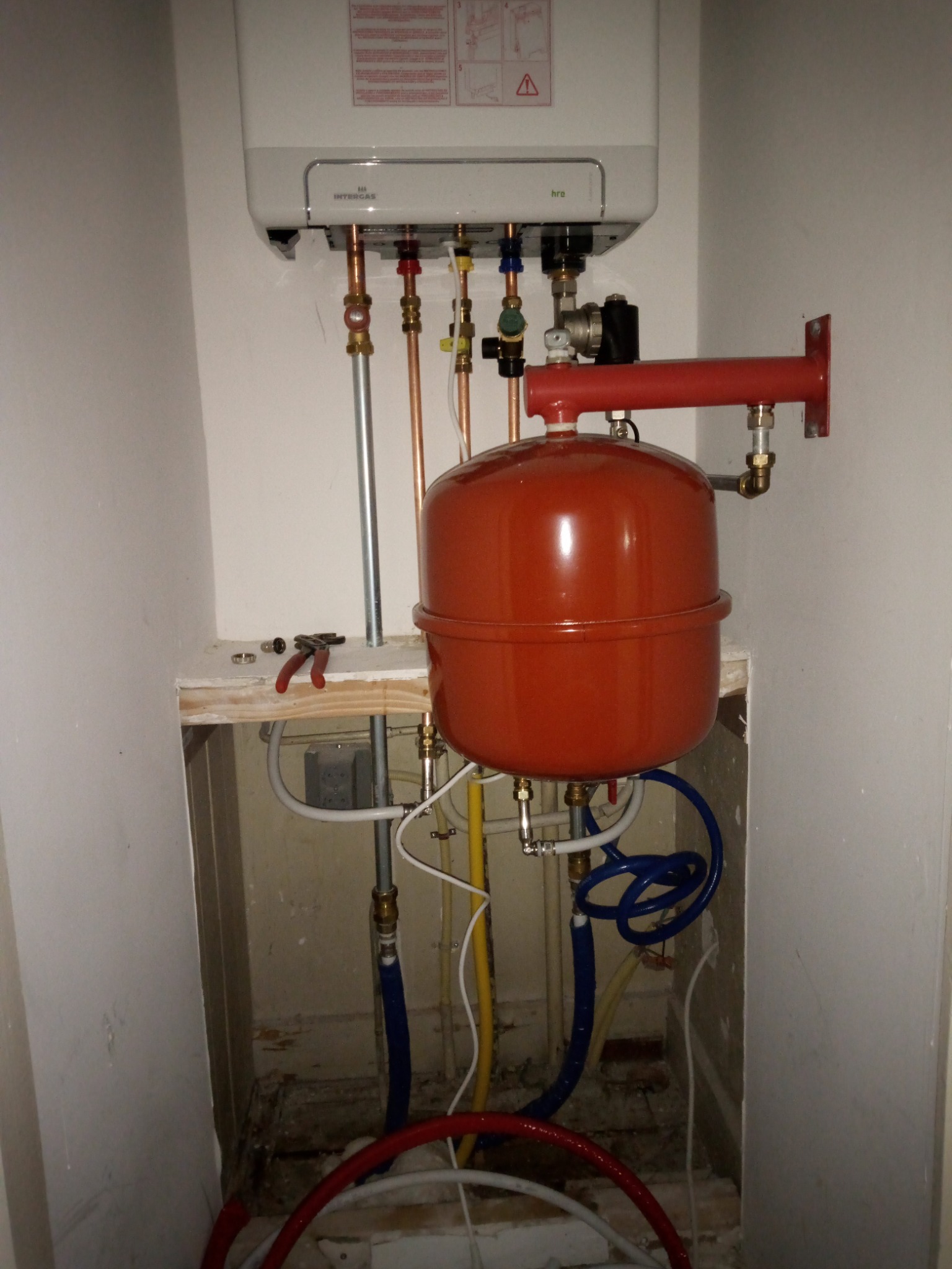 Biały piec gazowy Intergas HRE, z podłączonym czerwonym zbiornikiem wyrównawczym, widoczne miedziane rury i zawory, pod spodem plątanina przewodów hydraulicznych na tle surowej ściany.
