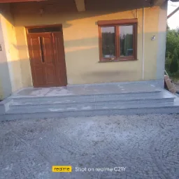 Trzy stopnie schodowe z jasnoszarego kamienia prowadzące do drzwi wejściowych domu, otoczone kostką brukową i zielenią.