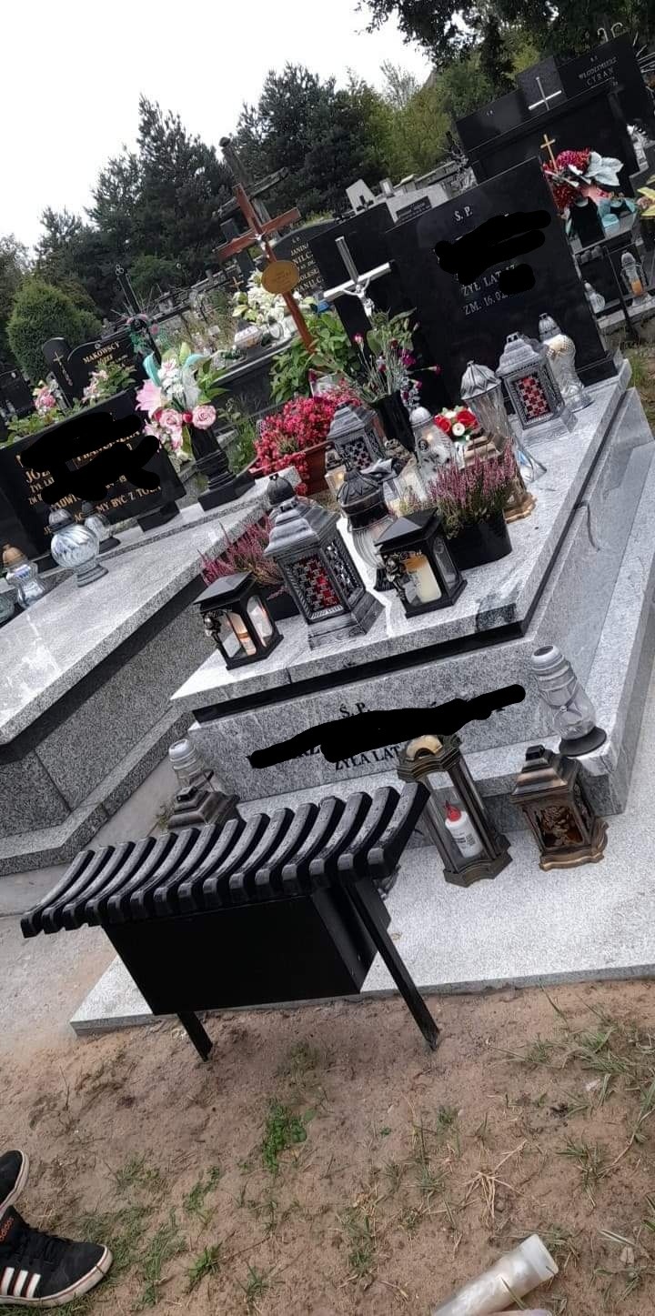 Widok na nagrobki z szarego kamienia z zapalonymi zniczami i kwiatami, w tle inne groby i drzewa, na pierwszym planie czarny metalowy kwietnik.