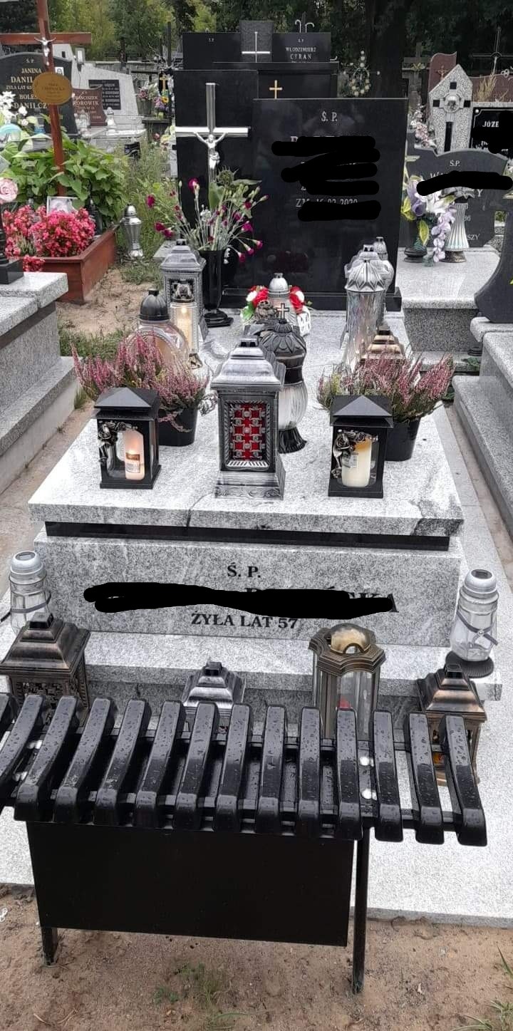 Granitowy pomnik nagrobny z zapalonymi lampionami, wrzosami w doniczkach i czarną ławeczką z metalowymi szczebelkami na cmentarzu.