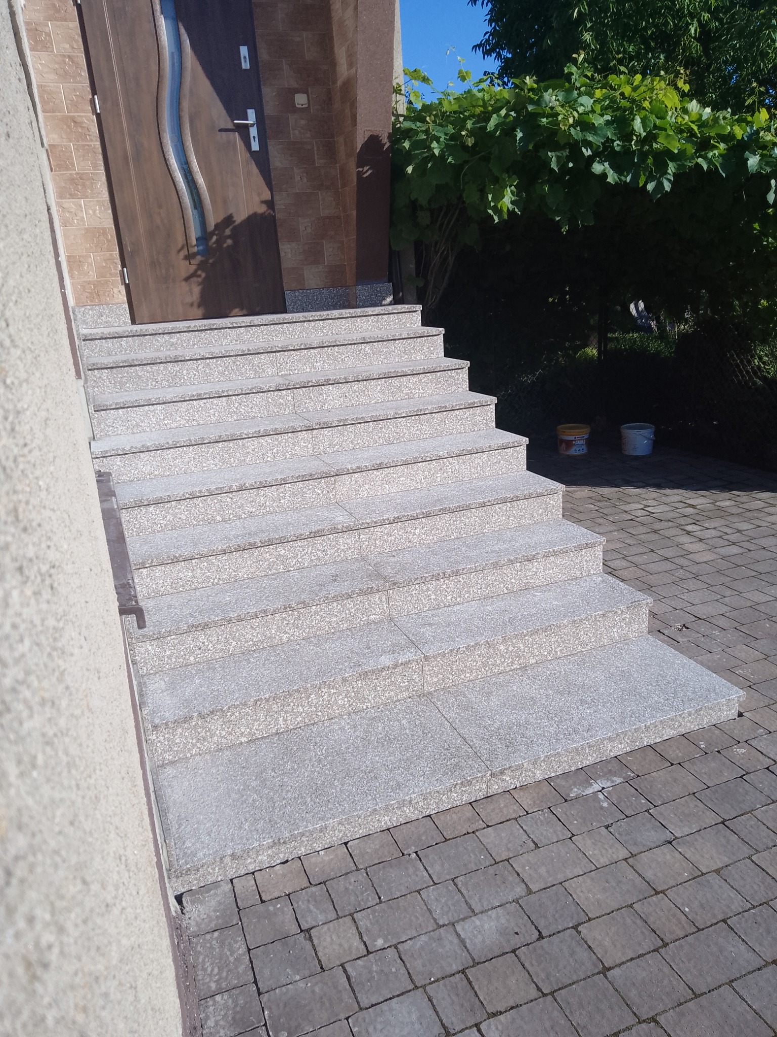 Zewnętrzne schody wejściowe z jasnego kamienia, prowadzące do drzwi wejściowych z dekoracyjnym szkłem, obok ściana budynku i kostka brukowa.