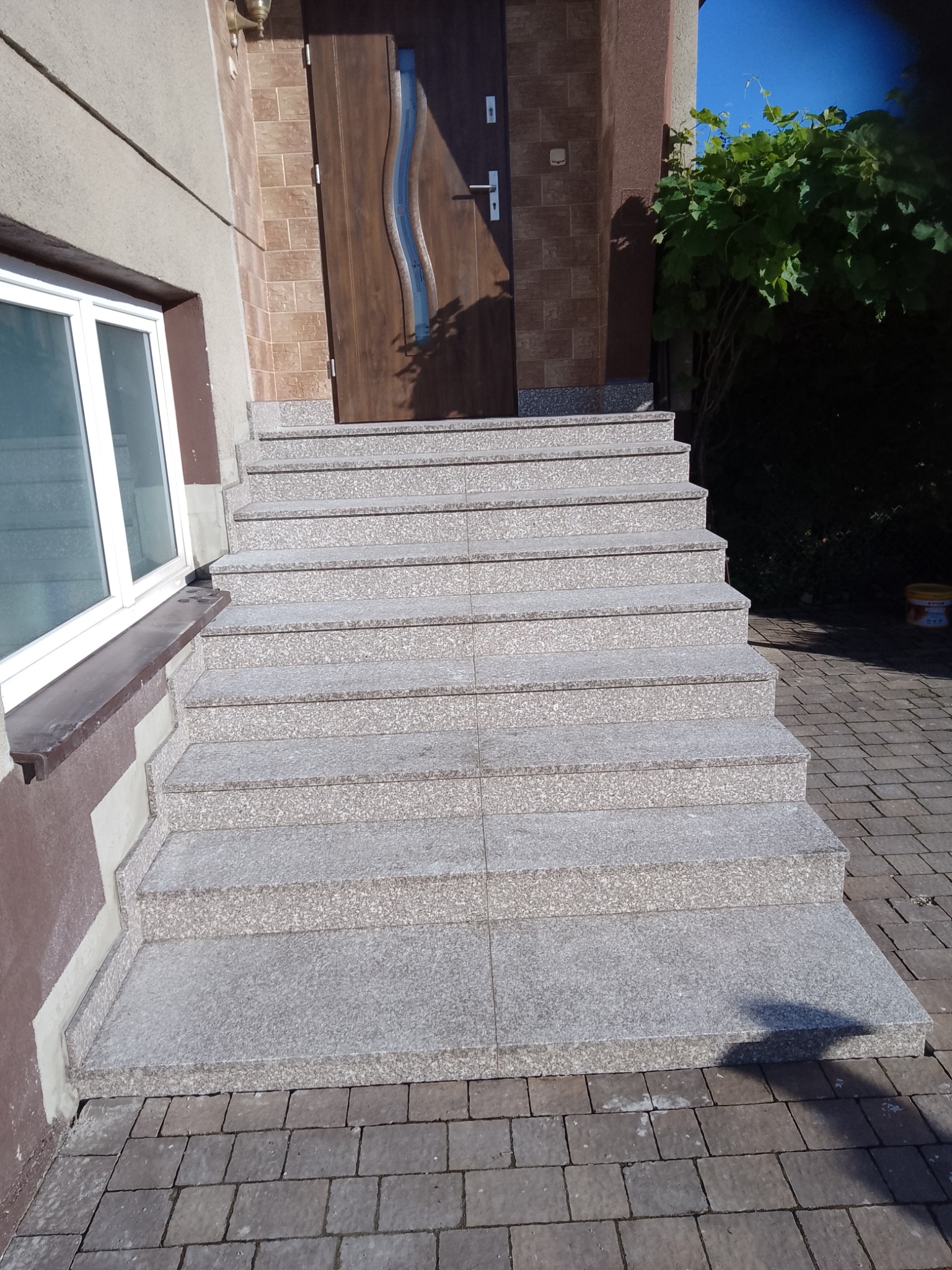 Zewnętrzne schody wejściowe wyłożone szarym kamieniem z widocznymi ośmioma stopniami, prowadzące do brązowych drzwi z dekoracyjnym przeszkleniem.