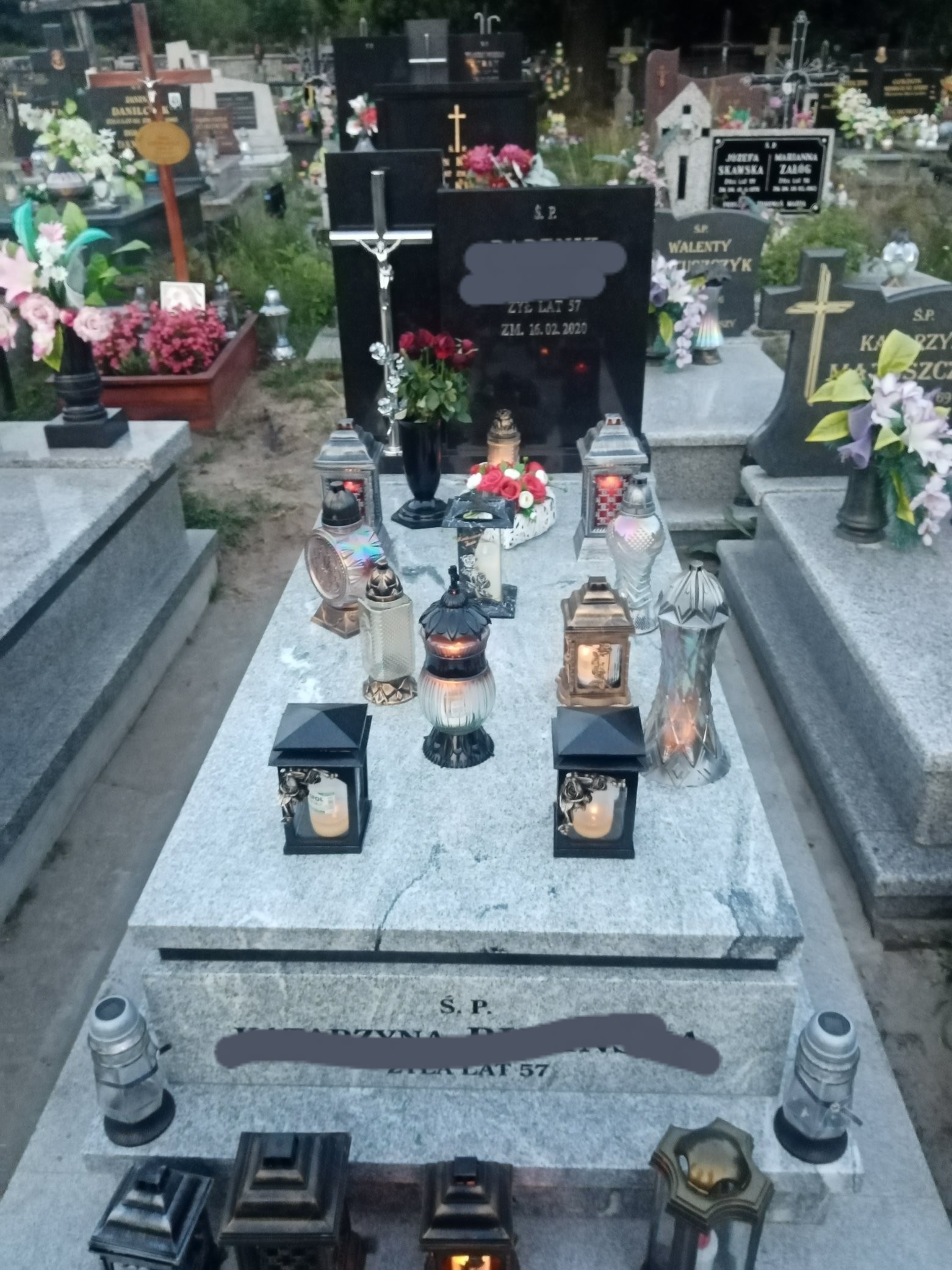 Granitowy pomnik z czarną tablicą, ozdobiony kwiatami, zniczami i lampionami na cmentarzu, widok z góry.