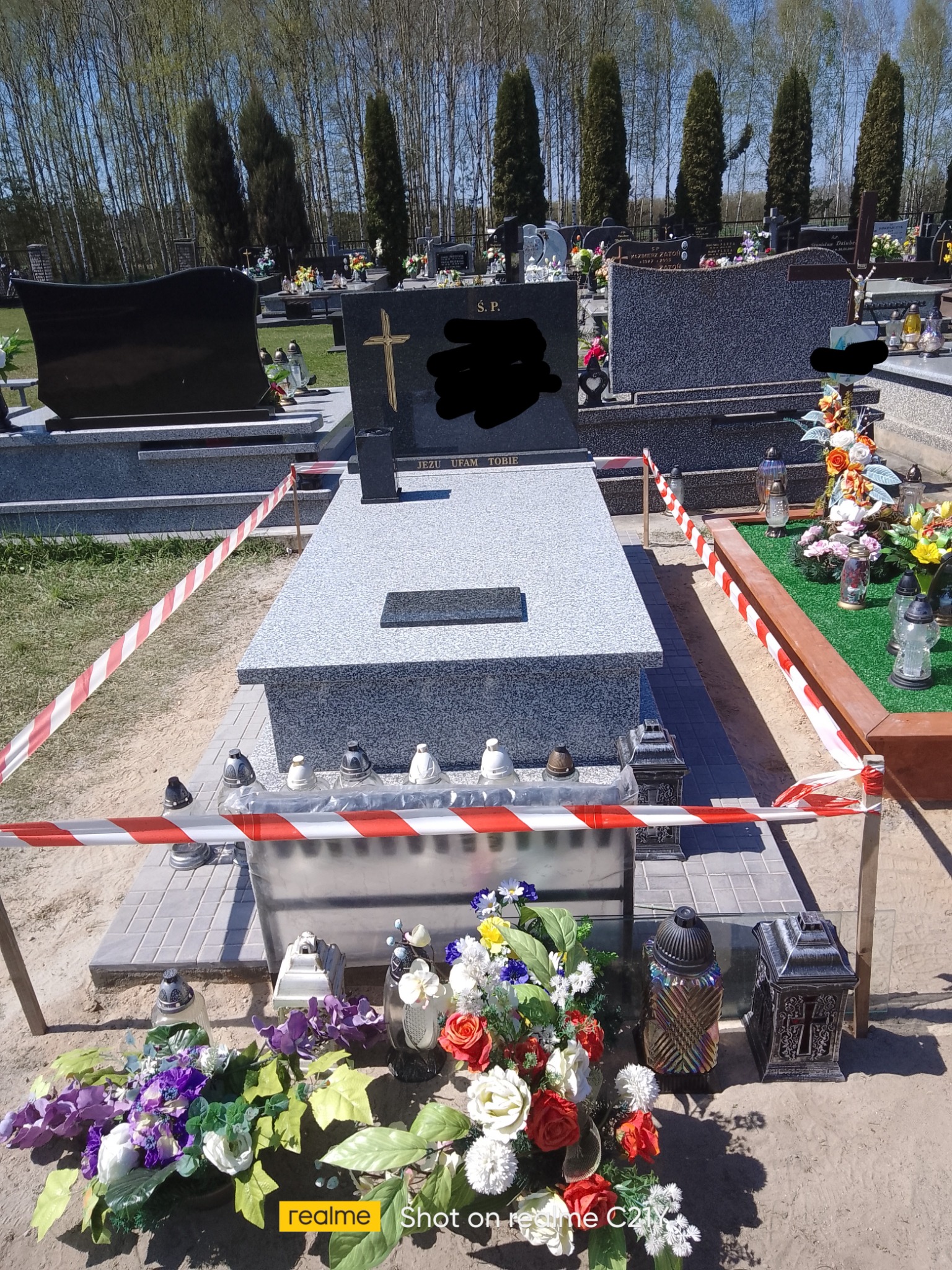 Szary nagrobek z jasnoszarą płytą, otoczony taśmą ostrzegawczą, z widocznymi zniczami i kwiatami na cmentarzu w słoneczny dzień.