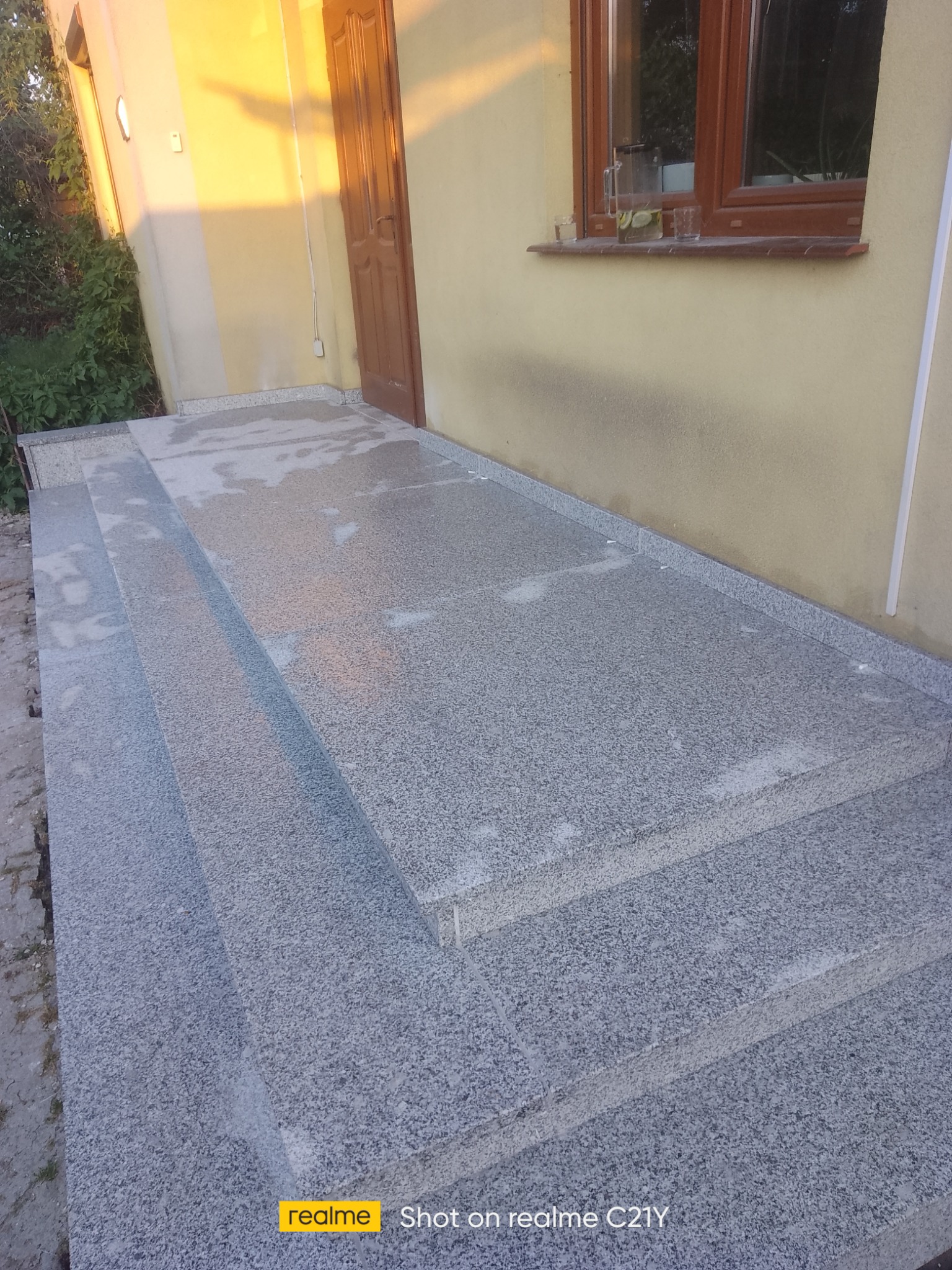 Zewnętrzne schody wejściowe wyłożone szarym, nakrapianym kamieniem, prowadzące do drzwi wejściowych domu z oknem, na którym stoi dzbanek z napojem.