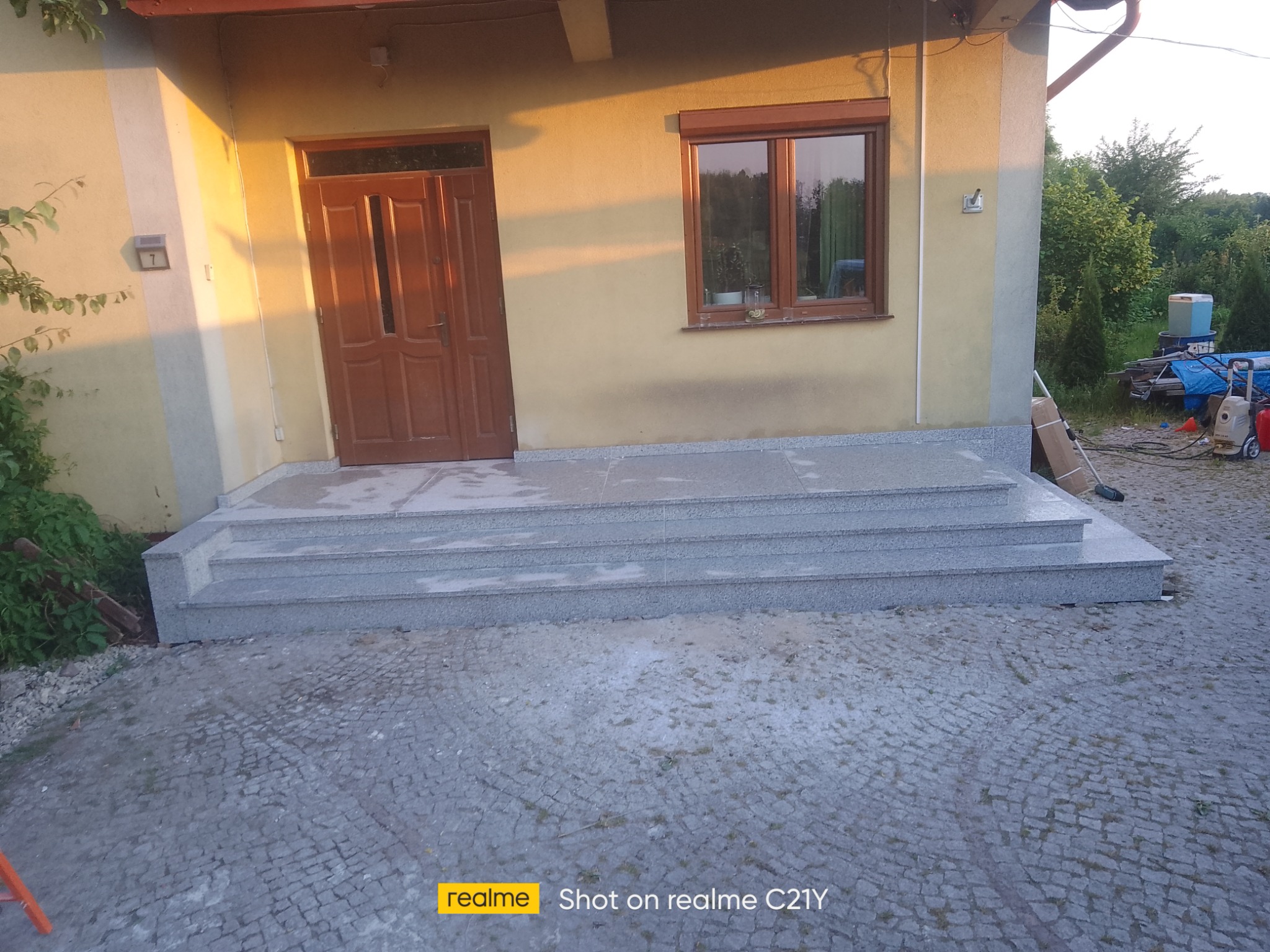 Trzy stopnie schodowe z jasnoszarego kamienia prowadzące do drzwi wejściowych domu, otoczone kostką brukową i zielenią.