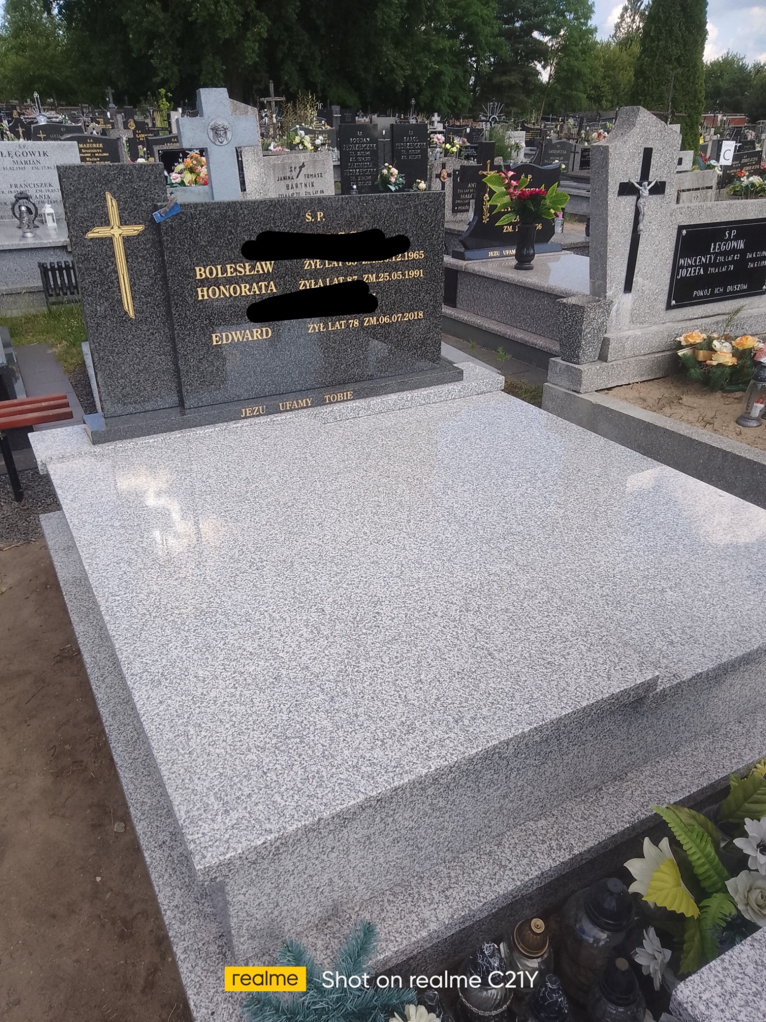 Szary, granitowy nagrobek z inskrypcjami i złotym krzyżem na tle cmentarza z innymi nagrobkami.