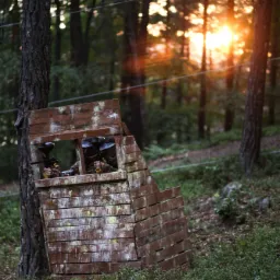 Dwie osoby z maskami i karabinami do paintballa ukryte w drewnianej fortyfikacji w lesie, promienie zachodzącego słońca w tle.