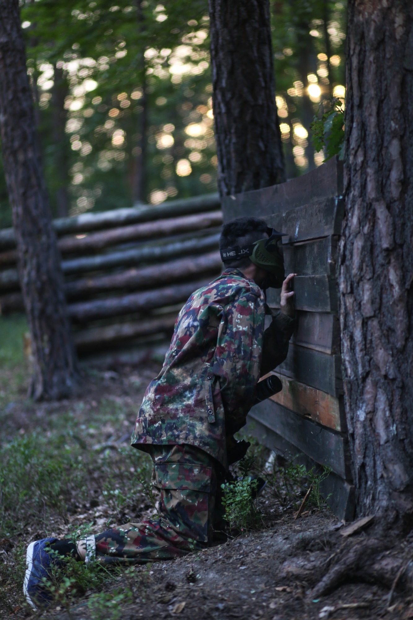 Osoba w kamuflażu, w masce ochronnej, przykuca za drewnianą przeszkodą podczas gry w paintball na zewnątrz, w tle widoczne drzewa i promienie słońca.
