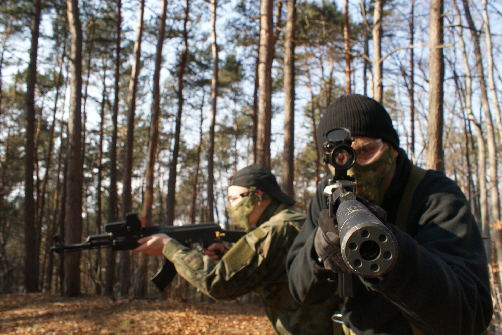 Dwóch zamaskowanych mężczyzn z replikami broni w lesie, jeden celuje prosto w obiektyw, widoczny przez lunetę, drugi w tle w kamuflażu.