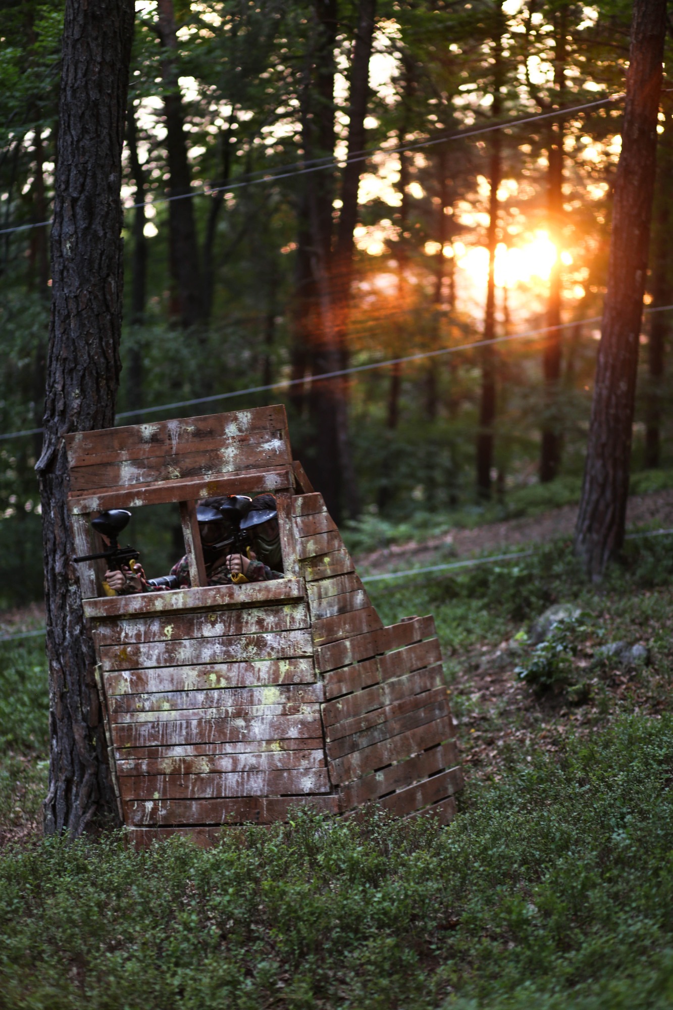 Dwie osoby z maskami i karabinami do paintballa ukryte w drewnianej fortyfikacji w lesie, promienie zachodzącego słońca w tle.