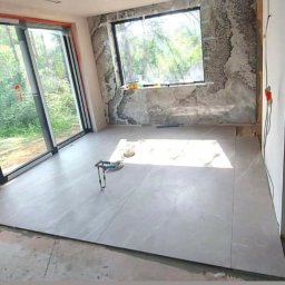 M&S usługi wykończeniowo/budowlane - Wnętrze w trakcie wykańczania: ułożone szare płytki podłogowe, częściowo wykończona ściana z efektem marmuru, duże okno z widokiem na zieleń, narzędzia na podłodze.