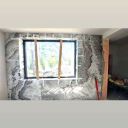 M&S usługi wykończeniowo/budowlane - Wnętrze w trakcie wykańczania ścian płytami imitującymi marmur, widoczne wsporniki montażowe i poziomica, okno z widokiem na zieleń.