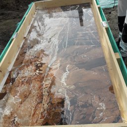 M&S usługi wykończeniowo/budowlane - Płyta z brązowego marmuru w drewnianej skrzyni transportowej, zabezpieczona folią, widoczny fragment ubrania roboczego osoby nadzorującej transport.