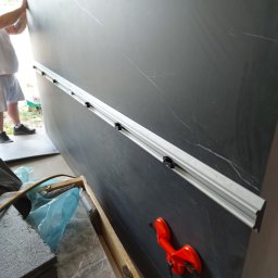 M&S usługi wykończeniowo/budowlane - Duża płyta z szarego marmuru z widocznym narzędziem do przenoszenia i listwą prowadzącą, przygotowywana do montażu.