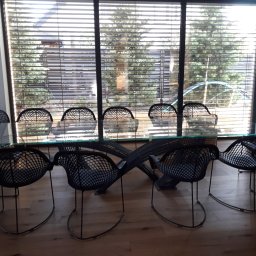 M&S usługi wykończeniowo/budowlane - Jadalnia z nowoczesnym stołem ze szklanym blatem i ażurowymi, czarnymi krzesłami na tle okna z żaluzjami, przez które widać ogród.