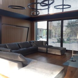 M&S usługi wykończeniowo/budowlane - Jasne, przestronne wnętrze w Wrocławiu z drewnianą podłogą, dużym narożnikiem w odcieniach szarości i nowoczesnym oświetleniem w postaci wiszących okręgów. Widoczne rusztowanie przy oknie sugeruje...