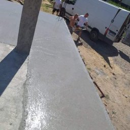 M&S usługi wykończeniowo/budowlane - Świeżo wylana, mokra, szara gładź na elewacji zewnętrznej budynku, z widocznymi śladami narzędzi i pracownikami w tle, zaparkowany biały van.