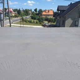 M&S usługi wykończeniowo/budowlane - Świeżo pomalowana elewacja budynku jednorodzinnego w odcieniach szarości, widok z bliska na gładką powierzchnię z delikatnymi śladami pędzla, w tle zabudowa mieszkalna i błękitne niebo.