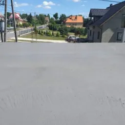 Świeżo pomalowana elewacja budynku jednorodzinnego w odcieniach szarości, widok z bliska na gładką powierzchnię z delikatnymi śladami pędzla, w tle zabudowa mieszkalna i błękitne niebo.