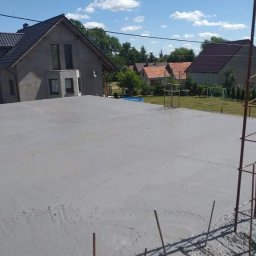 M&S usługi wykończeniowo/budowlane - Świeżo wylana, szara, betonowa wylewka na elewacji domu jednorodzinnego, widok z góry na tle zabudowy wiejskiej i błękitnego nieba.