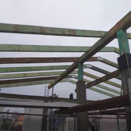 M&S usługi wykończeniowo/budowlane - Konstrukcja drewniana dachu w trakcie budowy, widoczne impregnowane belki i słupy, mocowania metalowe, tło stanowi szary mur i pochmurne niebo.