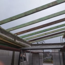 M&S usługi wykończeniowo/budowlane - Surowy stan konstrukcji dachu z drewnianymi belkami w kolorze zielonym i brązowym, widoczne metalowe łączniki, ściany z białych bloczków, szare niebo w tle.