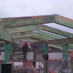 M&S usługi wykończeniowo/budowlane - Konstrukcja więźby dachowej z impregnowanego drewna, widoczny pracownik na rusztowaniu i drabina, w tle zabudowa jednorodzinna.