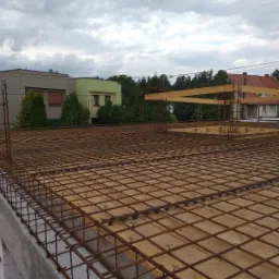 Zbrojenie płyty stropowej z widocznymi elementami dystansowymi i szalunkiem drewnianym, w tle zabudowa jednorodzinna i pochmurne niebo.