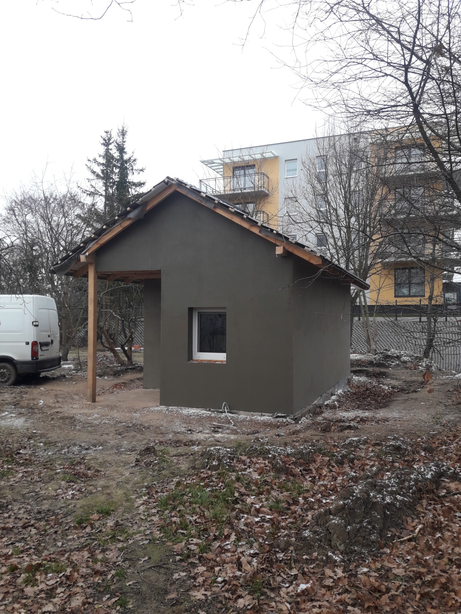 Surowy budynek z otynkowanymi ścianami zewnętrznymi, widoczny podbitka dachu i fragment białego samochodu dostawczego, na tle drzew i bloku mieszkalnego.