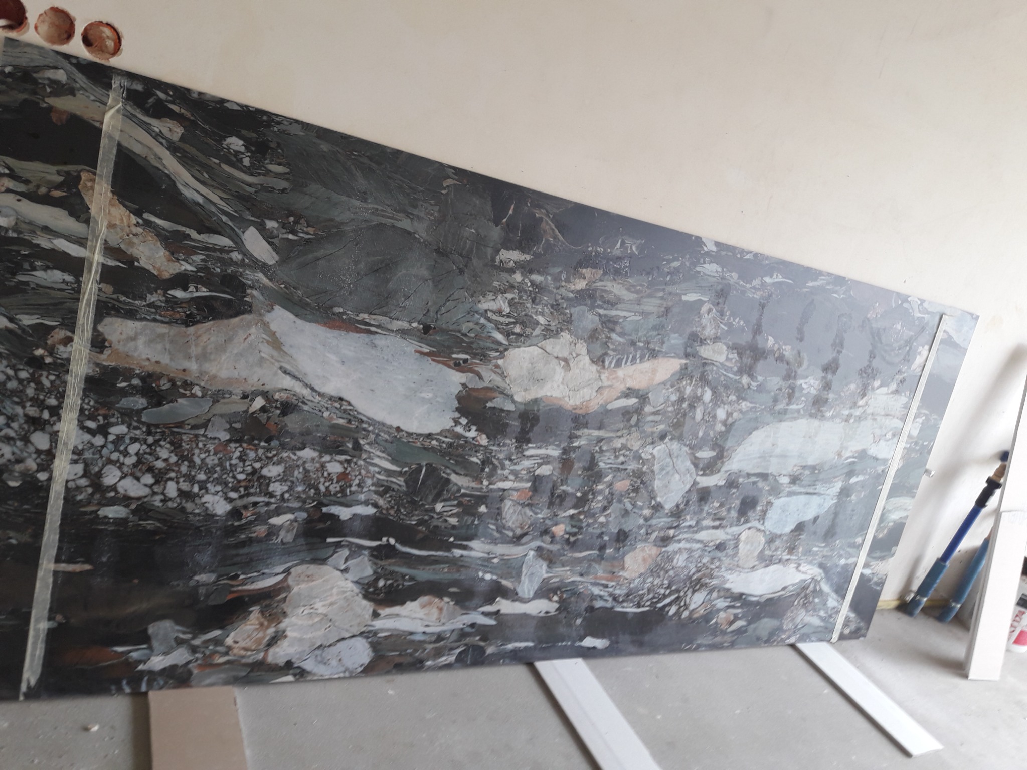 Duża płyta marmurowa z unikalnym wzorem, oparta o ścianę w trakcie montażu, widoczne otwory w ścianie i narzędzia w tle.