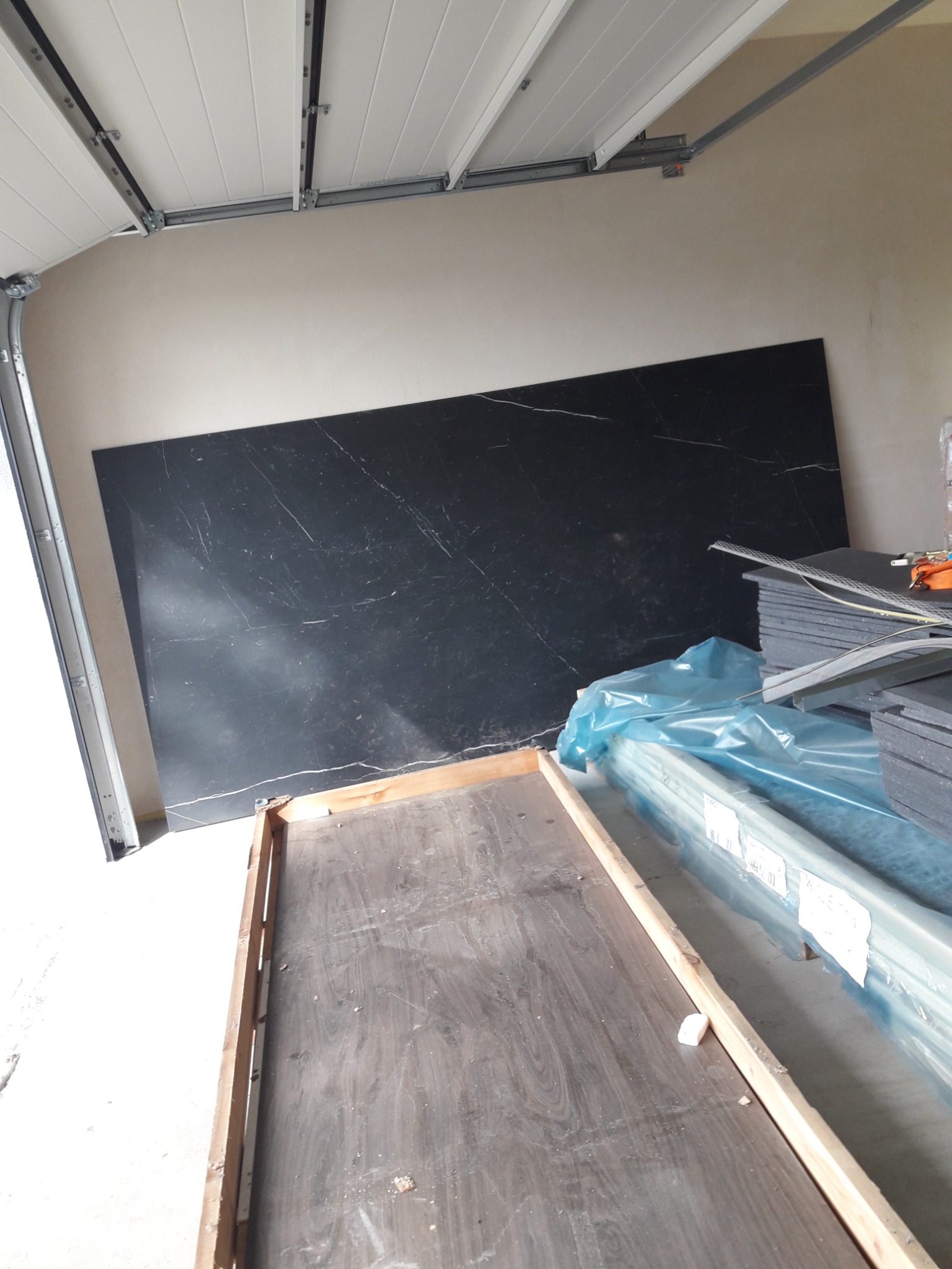 Duża płyta z czarnego marmuru z białymi żyłami oparta o ścianę w garażu obok materiałów budowlanych w folii i drewnianej ramy.