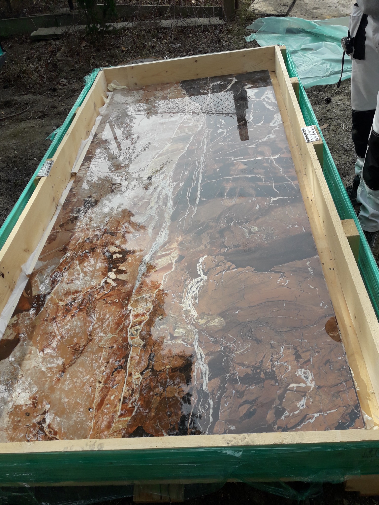 Płyta z brązowego marmuru w drewnianej skrzyni transportowej, zabezpieczona folią, widoczny fragment ubrania roboczego osoby nadzorującej transport.
