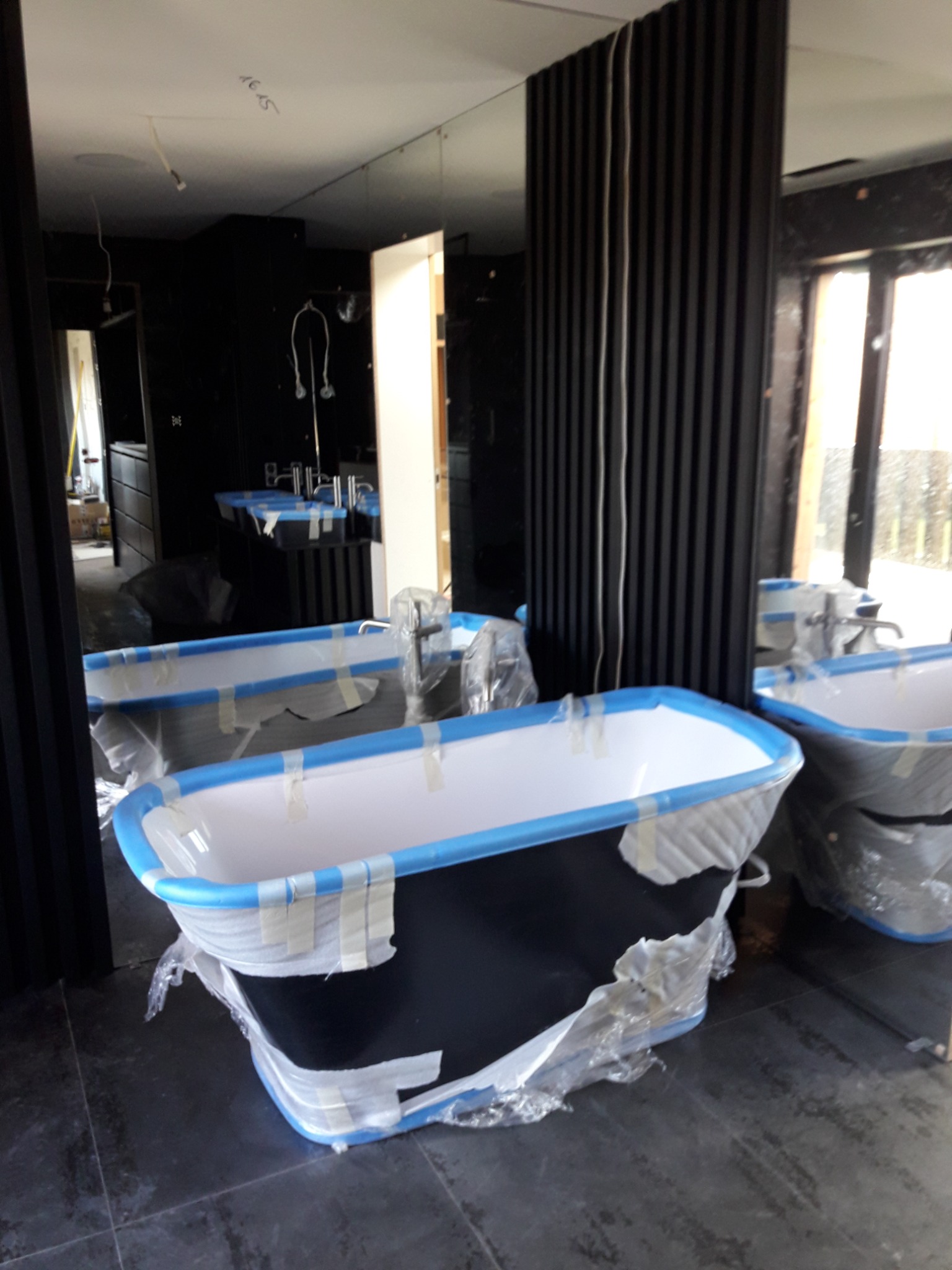 Nowa, czarna wanna wolnostojąca zabezpieczona folią i taśmą malarską w trakcie remontu łazienki z ciemnymi płytkami i elementami dekoracyjnymi z pionowych listew.