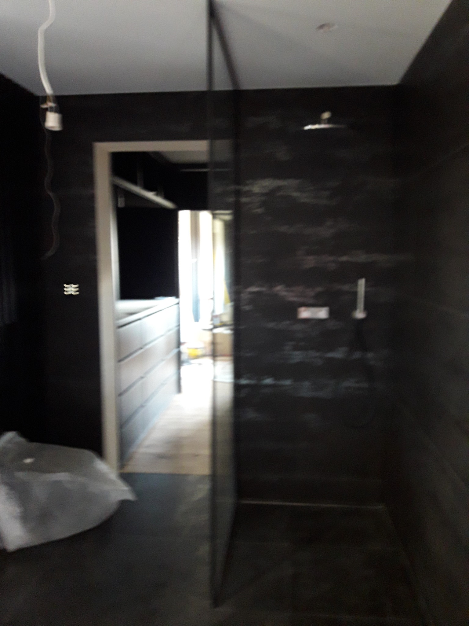 Ciemna łazienka z widoczną kabiną prysznicową i wejściem do garderoby, częściowo zasłonięta folią ochronną na podłodze podczas remontu.