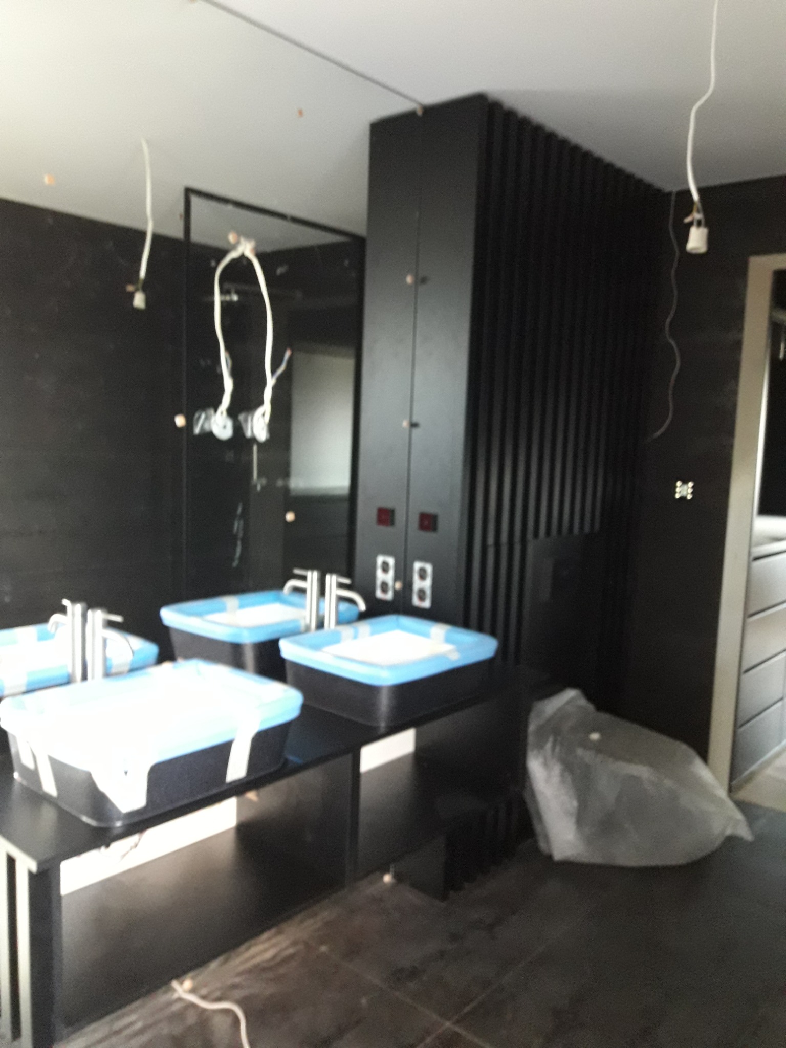 Niedokończona łazienka z trzema umywalkami w czarnych obudowach z niebieskimi akcentami, widoczne wiszące przewody elektryczne i zabezpieczone elementy wyposażenia.
