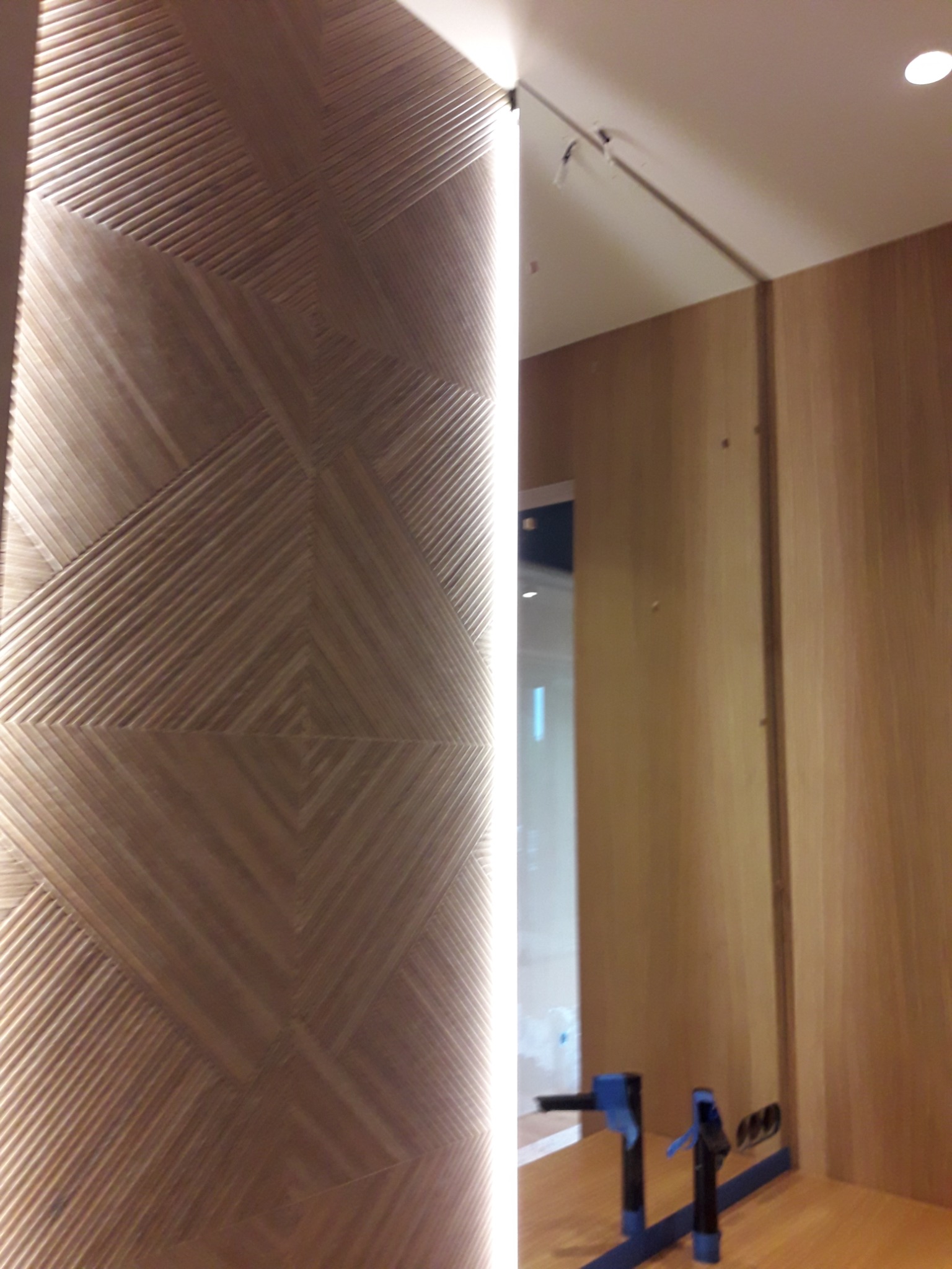Ściana z dekoracyjną drewnianą okładziną w geometryczny wzór, oświetlona pionowym pasem światła LED, widoczne narzędzia montażowe.