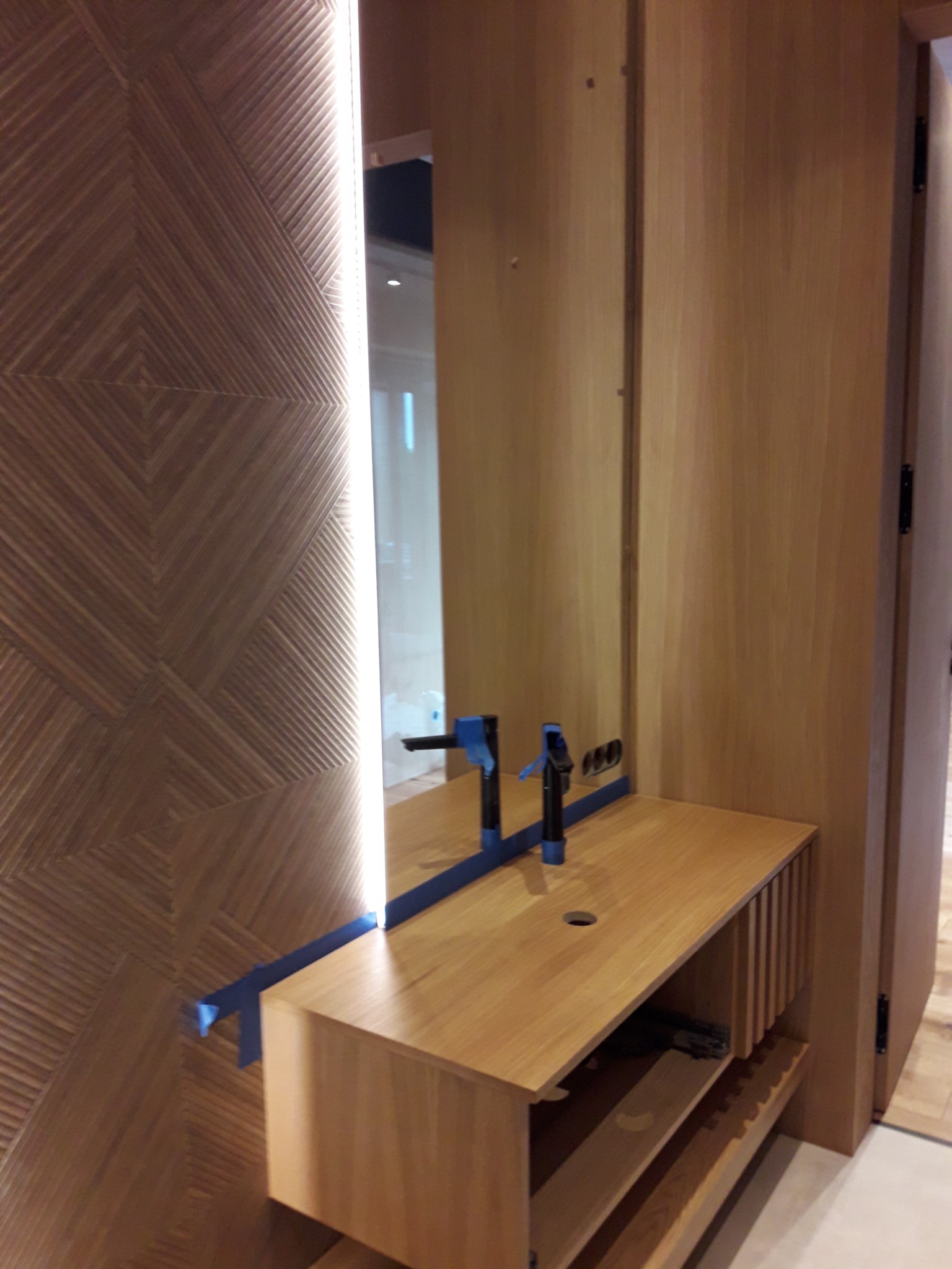 Montaż baterii umywalkowej na drewnianym blacie z podświetlanym lustrem i dekoracyjną ścianą z drewnianych paneli, fragment łazienki w trakcie remontu.