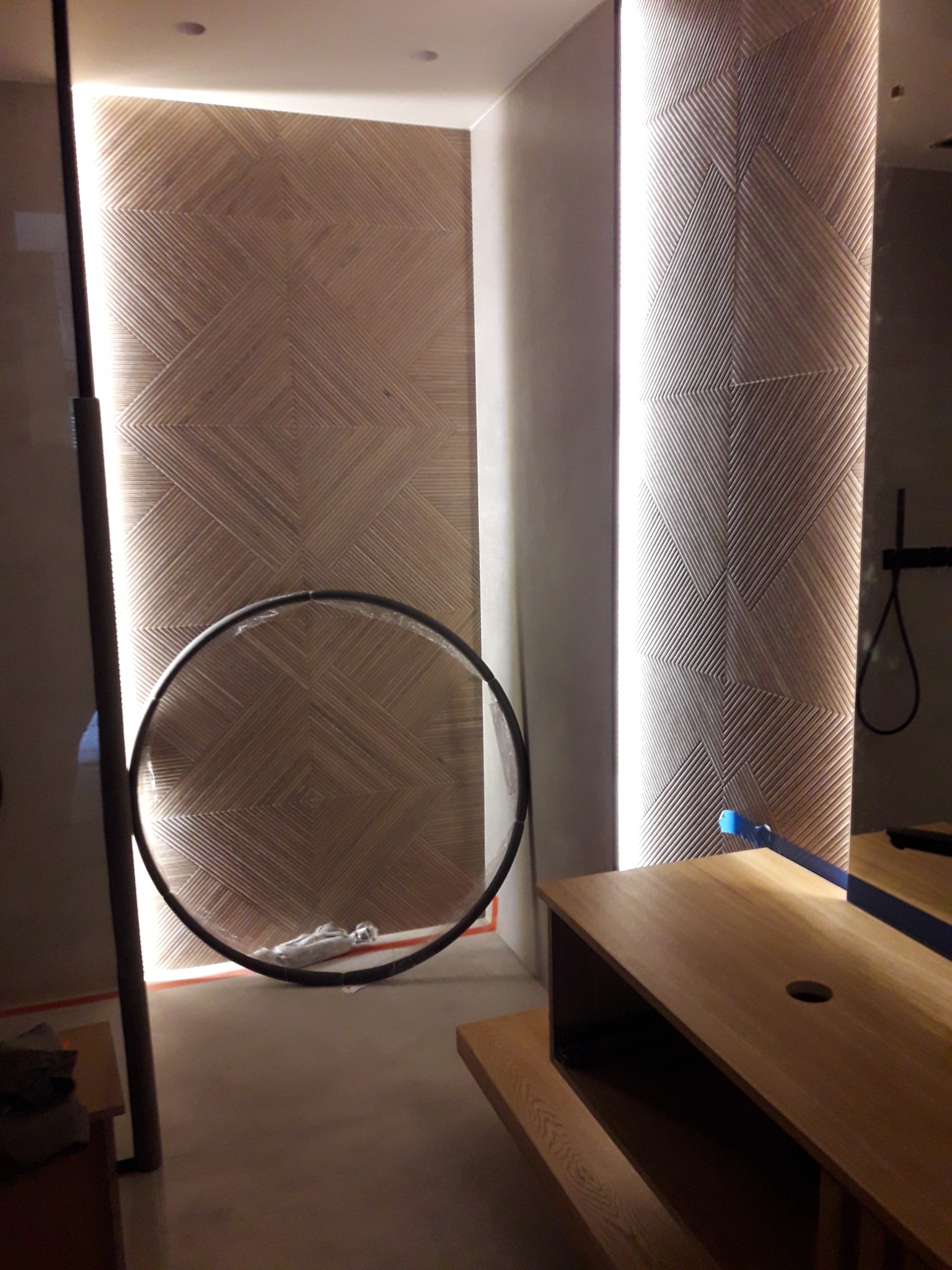 Ściana z dekoracyjną strukturą, podświetlona listwami LED, z okrągłym elementem dekoracyjnym i fragmentem drewnianego mebla w minimalistycznym wnętrzu.