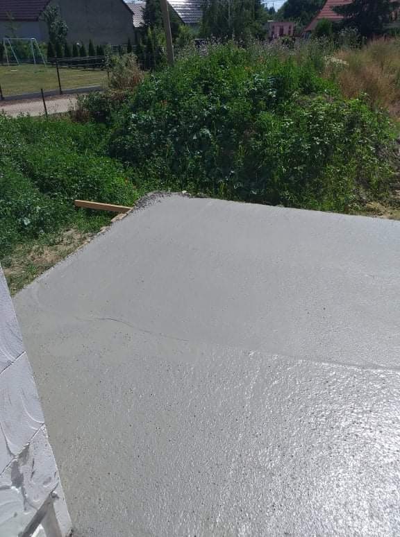 Świeżo wylana, mokra, szara wylewka betonowa na zewnątrz budynku, z widocznymi śladami wyrównywania, obok ściana z białego bloczka.