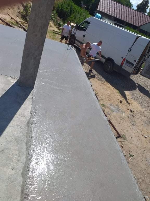 Świeżo wylana, mokra, szara gładź na elewacji zewnętrznej budynku, z widocznymi śladami narzędzi i pracownikami w tle, zaparkowany biały van.