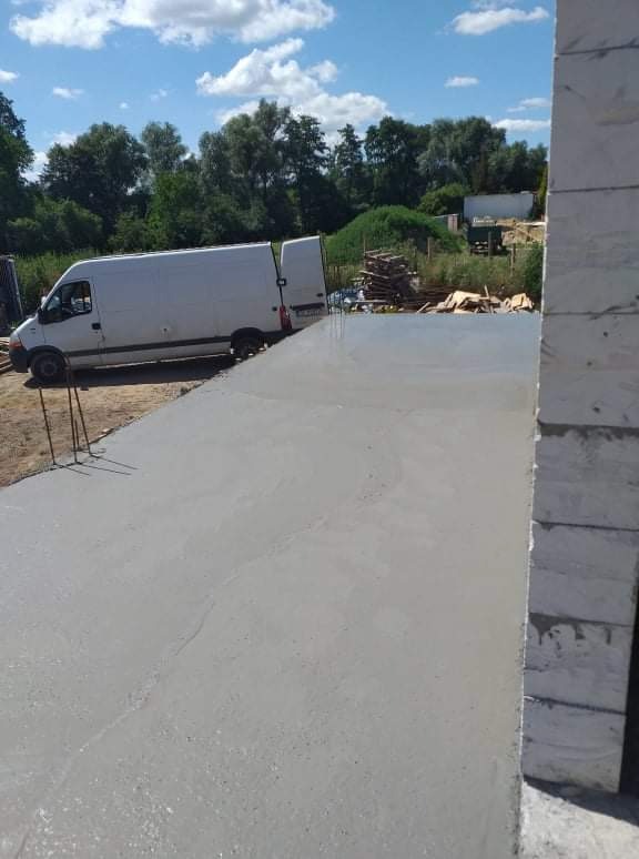 Świeżo wylana, gładka posadzka betonowa na zewnątrz budynku we Wrocławiu, z widocznym samochodem dostawczym w tle i stosem desek.