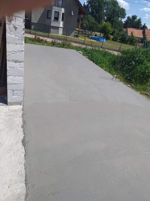 Świeżo wylana, gładka betonowa wylewka na podjeździe, widoczny fragment ściany budynku z białej cegły i zieleń w tle.