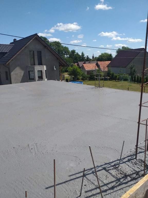 Świeżo wylana, szara, betonowa wylewka na elewacji domu jednorodzinnego, widok z góry na tle zabudowy wiejskiej i błękitnego nieba.