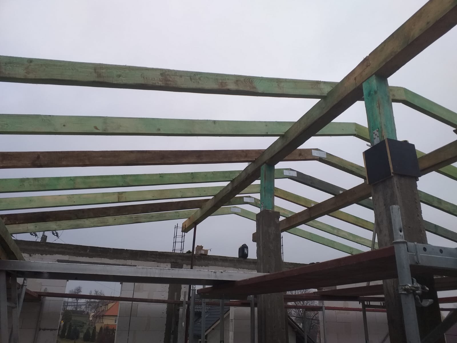 Konstrukcja drewniana dachu w trakcie budowy, widoczne impregnowane belki i słupy, mocowania metalowe, tło stanowi szary mur i pochmurne niebo.