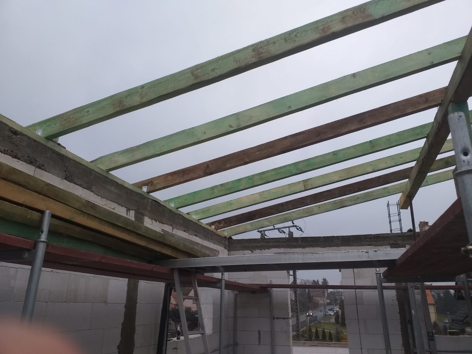 Surowy stan konstrukcji dachu z drewnianymi belkami w kolorze zielonym i brązowym, widoczne metalowe łączniki, ściany z białych bloczków, szare niebo w tle.