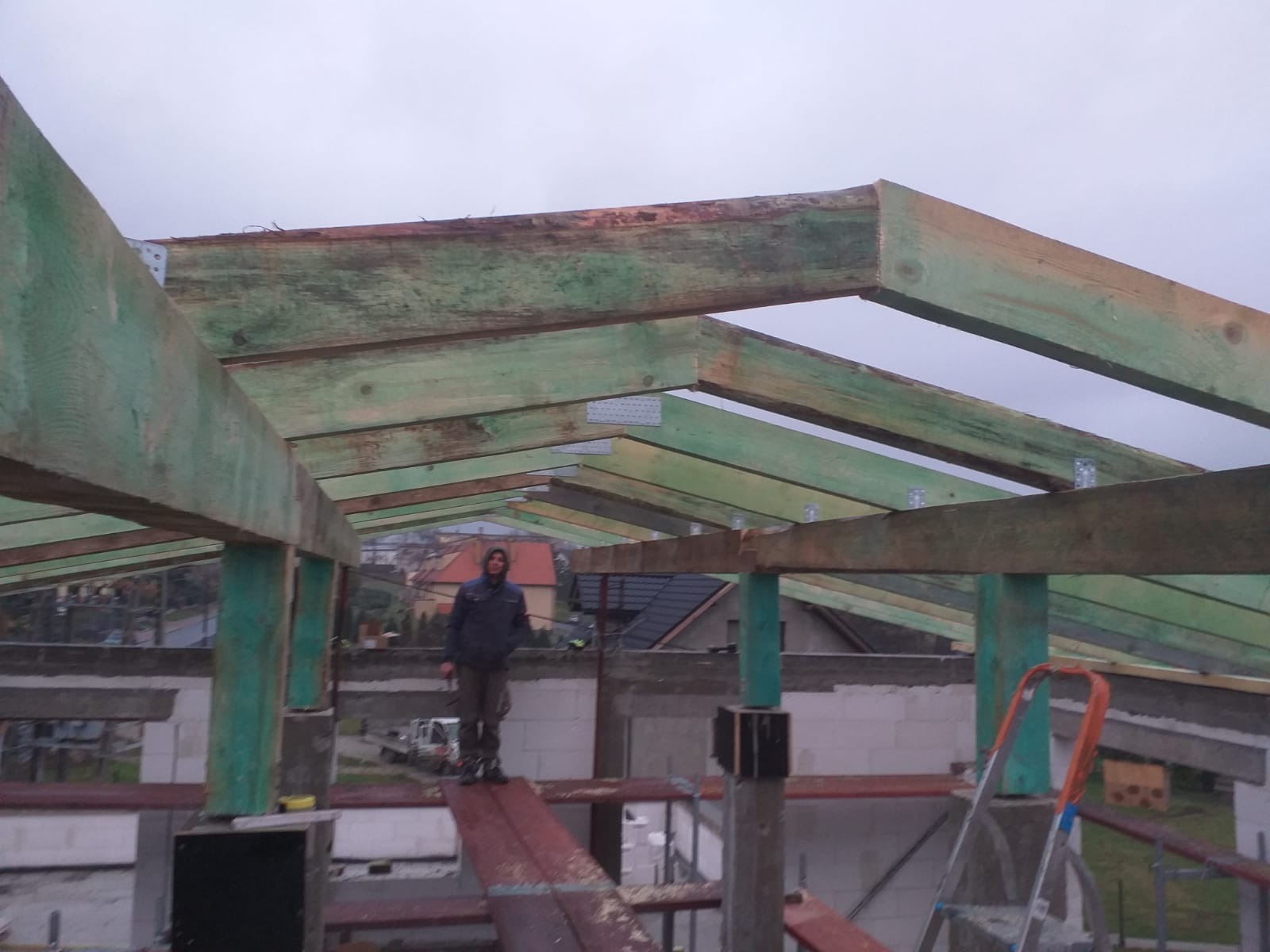 Konstrukcja więźby dachowej z impregnowanego drewna, widoczny pracownik na rusztowaniu i drabina, w tle zabudowa jednorodzinna.